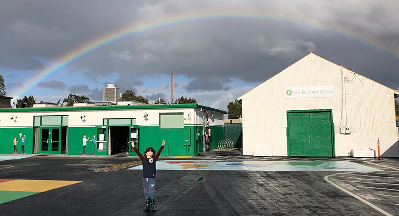 Person on a playground with a rainbow in the distance over school buildings