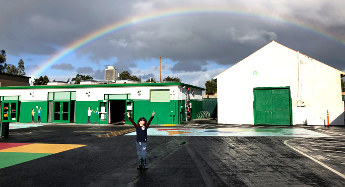 Person on a playground with a rainbow in the distance over school buildings